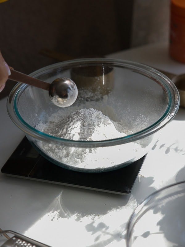 Easy Vegan Lemon Cookies Recipe (Soft and Lemony!) 14 A glass bowl filled with flour sits on a digital kitchen scale. A hand holds a metal measuring spoon above the bowl, ready to add an ingredient for lemon cookies. Sunlight casts shadows on the white surface beneath.