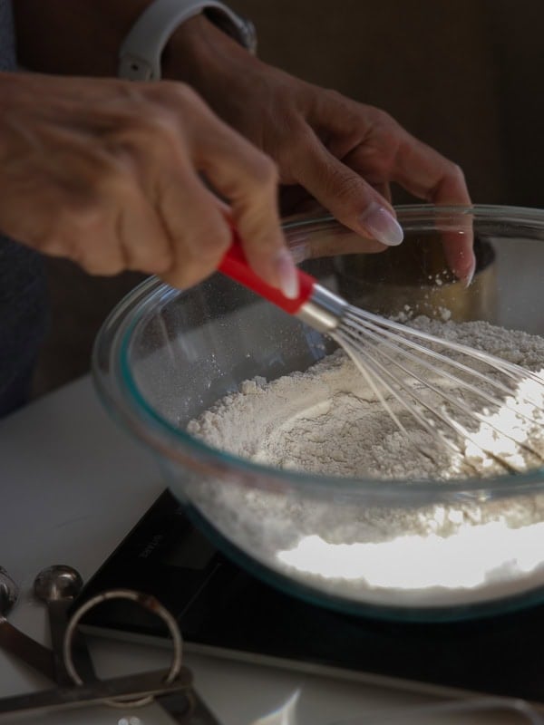 Easy Vegan Lemon Cookies Recipe (Soft and Lemony!) 15 A person uses a red-handled whisk to mix flour in a glass bowl, preparing dough for lemon cookies. Measuring spoons and kitchen tools are nearby on a white surface.