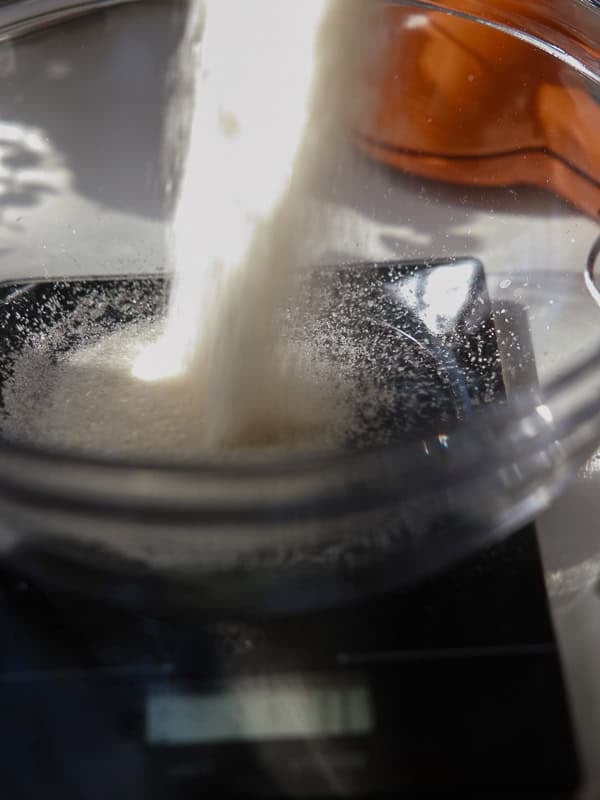 Easy Vegan Lemon Cookies Recipe (Soft and Lemony!) 5 A close-up of sugar being poured into a glass bowl on a digital kitchen scale, with sunlight illuminating the motionโperfect for making lemon cookies. A blurred orange object sits in the background.