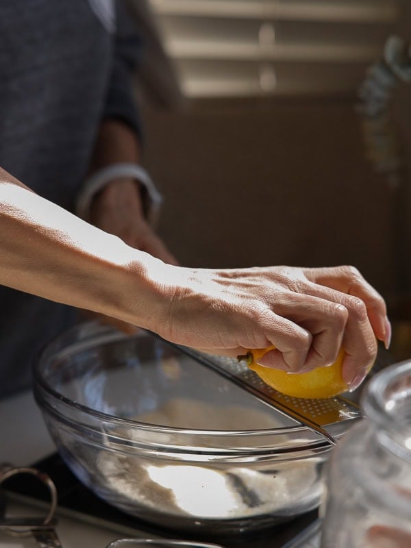 Easy Vegan Lemon Cookies Recipe (Soft and Lemony!) 6 A person zesting a lemon over a glass mixing bowl filled with flour, using a grater, in a sunlit kitchen while preparing lemon cookies.