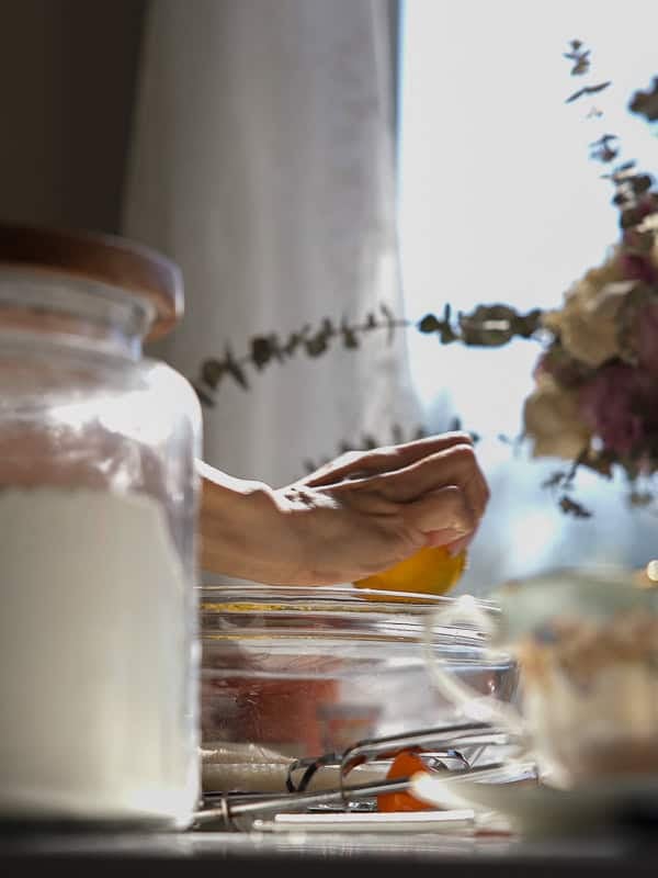 Easy Vegan Lemon Cookies Recipe (Soft and Lemony!) 3 A hand zests a lemon into a glass mixing bowl near a jar of flour, sunlight pouring in as lemon cookies are prepared, with a vase of flowers in the background.