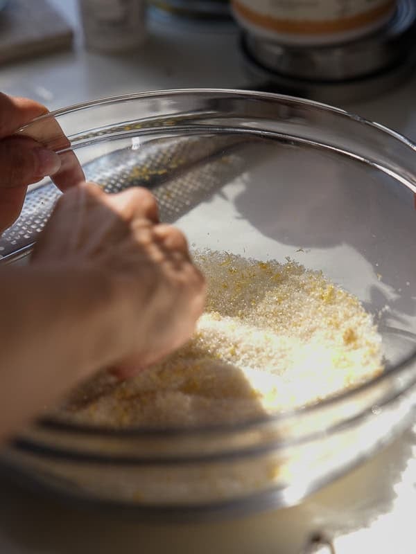 Easy Vegan Lemon Cookies Recipe (Soft and Lemony!) 8 A close-up of hands grating lemon zest into a glass bowl filled with sugar for homemade lemon cookies, with sunlight shining on the mixture and a grater visible inside the bowl.