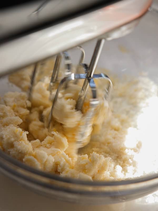 Easy Vegan Lemon Cookies Recipe (Soft and Lemony!) 10 Close-up of an electric hand mixer blending butter and sugar in a glass bowl for lemon cookies, creating a creamy mixture. The metal beaters are in motion, with the ingredients appearing partially mixed.