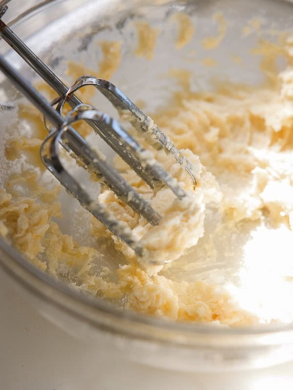 Easy Vegan Lemon Cookies Recipe (Soft and Lemony!) 11 A hand mixer with two beaters is mixing a creamy, pale lemon cookie dough in a clear glass bowl, with sunlight illuminating part of the mixture.