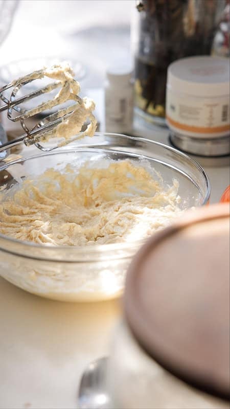 Easy Vegan Lemon Cookies Recipe (Soft and Lemony!) 13 A glass bowl filled with creamy lemon cookie batter sits on a kitchen counter. An electric mixer with batter-coated beaters rests above the bowl, while various jars and containers are in the background.