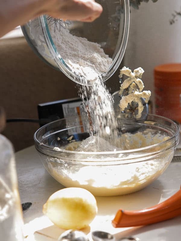 Easy Vegan Lemon Cookies Recipe (Soft and Lemony!) 16 A hand pours flour from a glass bowl into another bowl of batter on a kitchen counter. A lemon hints at lemon cookies in the making, while measuring spoons and baking tools rest nearby. Sunlight illuminates the scene.