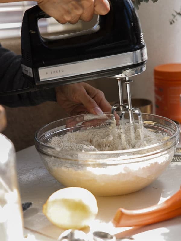 Easy Vegan Lemon Cookies Recipe (Soft and Lemony!) 17 A person uses a black electric hand mixer to blend ingredients in a glass bowl for lemon cookies; flour is visible on top of the mixture, with a lemon and measuring spoons nearby on a sunlit white surface.