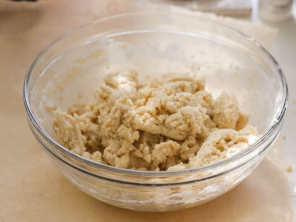 Easy Vegan Lemon Cookies Recipe (Soft and Lemony!) 18 A clear glass bowl filled with rough, partially mixed lemon cookie dough sits on a light-colored surface under bright natural light.