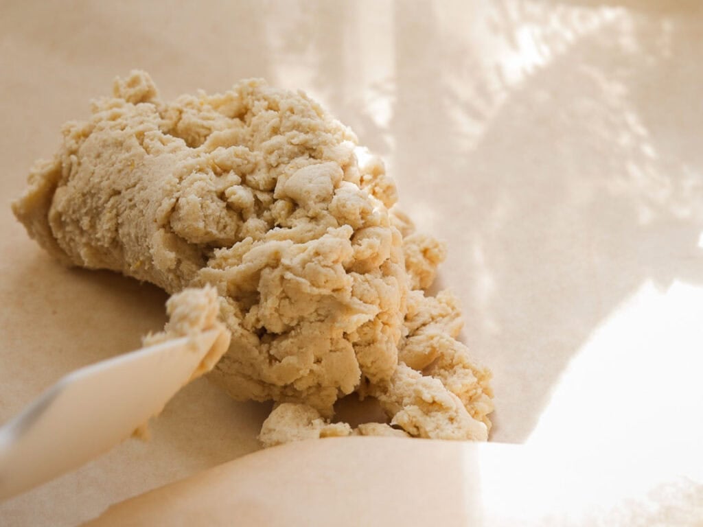 Easy Vegan Lemon Cookies Recipe (Soft and Lemony!) 19 A close-up of a ball of raw dough for lemon cookies on parchment paper, with a spatula next to it. Sunlight and shadows fall across the surface.
