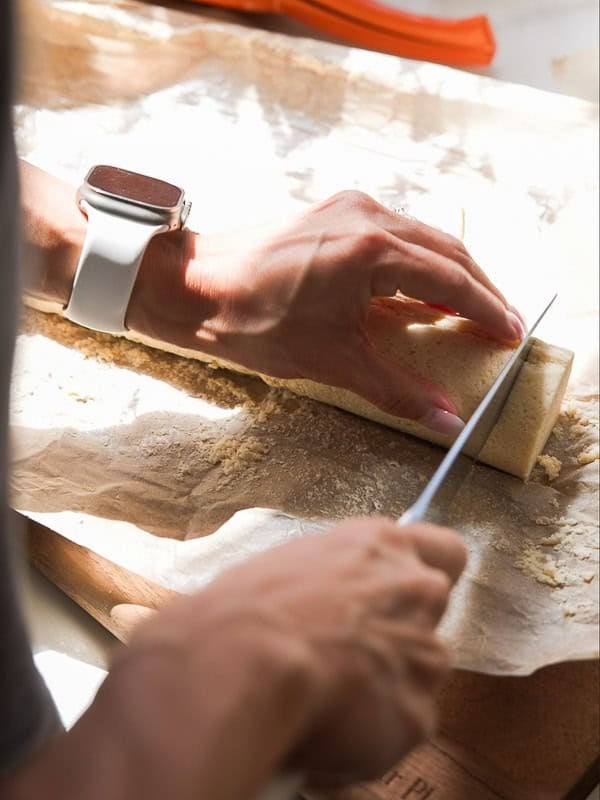 Easy Vegan Lemon Cookies Recipe (Soft and Lemony!) 21 A person wearing a white smartwatch slices a rolled lemon cookies dough log on parchment paper with a knife, preparing to bake. Sunlight highlights their hands and the work surface.