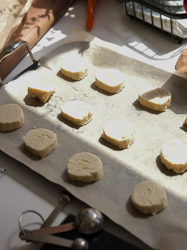 Easy Vegan Lemon Cookies Recipe (Soft and Lemony!) 22 A baking tray lined with parchment paper holds twelve unbaked lemon cookie dough slices. Measuring spoons and sunlight are visible on the white countertop nearby.