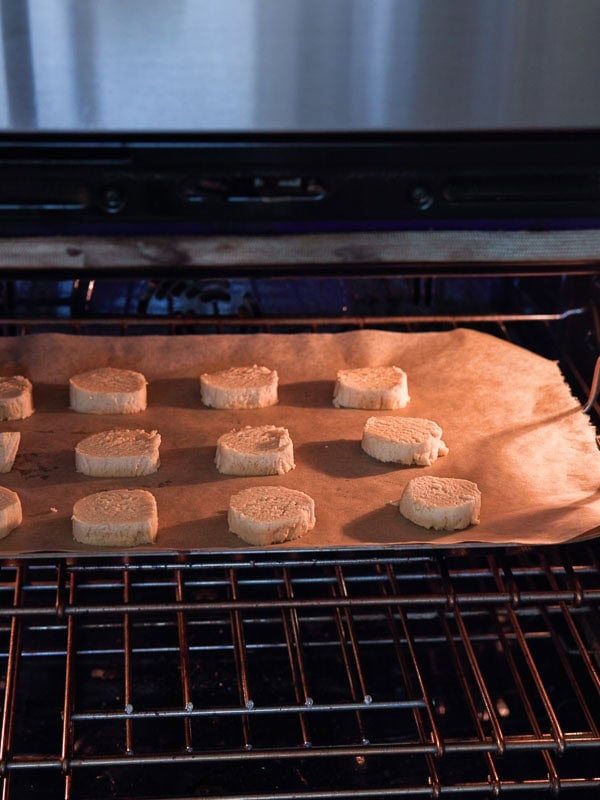 Easy Vegan Lemon Cookies Recipe (Soft and Lemony!) 23 Round slices of lemon cookie dough are spaced out on a parchment-lined baking sheet inside an oven, beginning to bake.