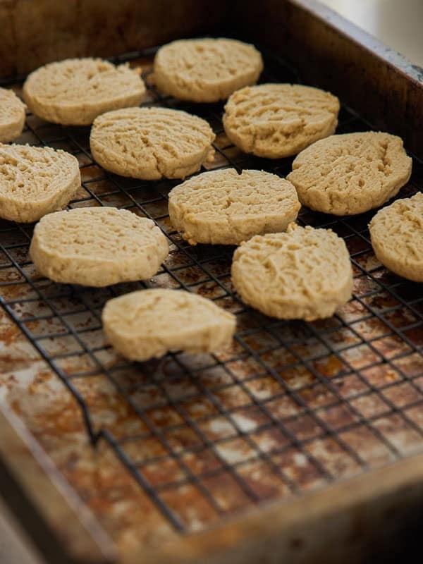 Easy Vegan Lemon Cookies Recipe (Soft and Lemony!) 4 Round, golden lemon cookies are cooling on a black wire rack placed inside a worn, metal baking pan. The cookies have a soft, slightly crumbly texture and are spaced apart from each other.