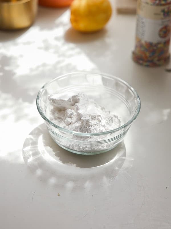 Easy Vegan Lemon Cookies Recipe (Soft and Lemony!) 25 A clear glass bowl filled with white powdered sugar sits on a sunlit white surface, ready to dust freshly baked lemon cookies. Shadow patterns and blurred colorful objects appear in the background.