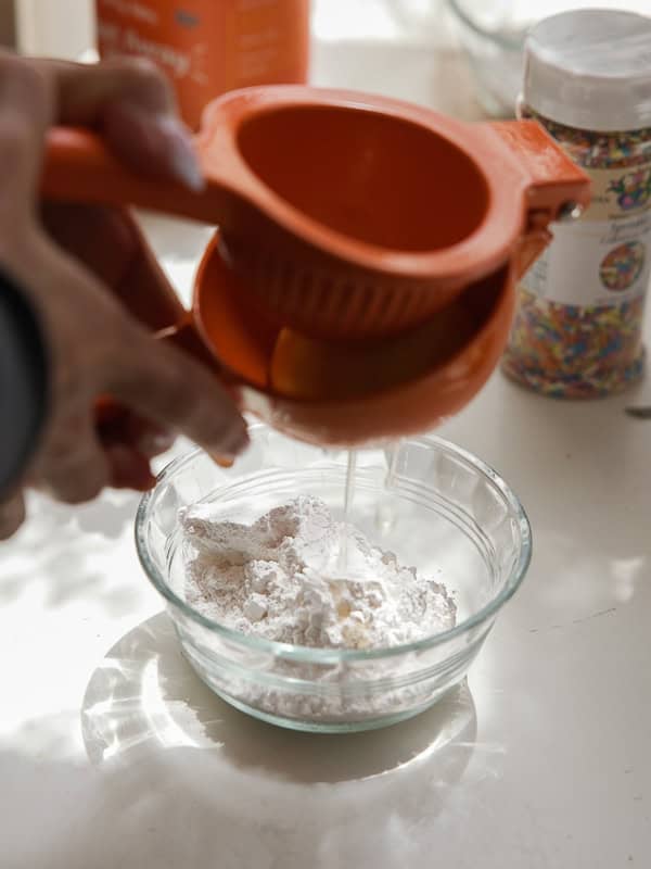 Easy Vegan Lemon Cookies Recipe (Soft and Lemony!) 26 A hand squeezes a lemon with an orange citrus juicer over a glass bowl filled with powdered sugar, preparing the sweet glaze for homemade lemon cookies. Colorful sprinkles and baking ingredients decorate the background.