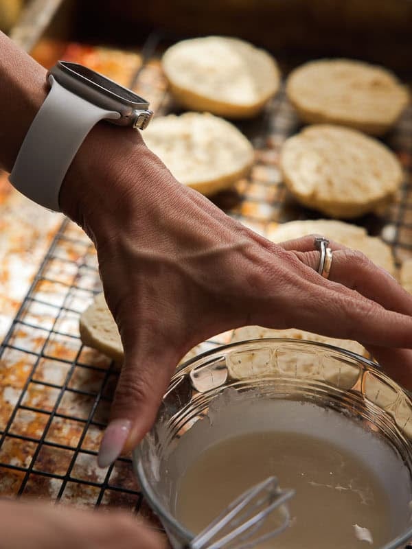 Easy Vegan Lemon Cookies Recipe (Soft and Lemony!) 27 A hand wearing a white watch and a wedding ring holds a small whisk in a glass bowl of icing, with freshly baked lemon cookies cooling on a wire rack in the background.