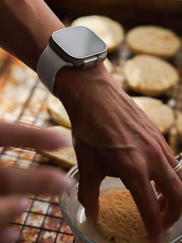 Easy Vegan Lemon Cookies Recipe (Soft and Lemony!) 28 A person wearing a smartwatch dips a biscuit into a bowl, with freshly baked lemon cookies cooling on a wire rack in the background.