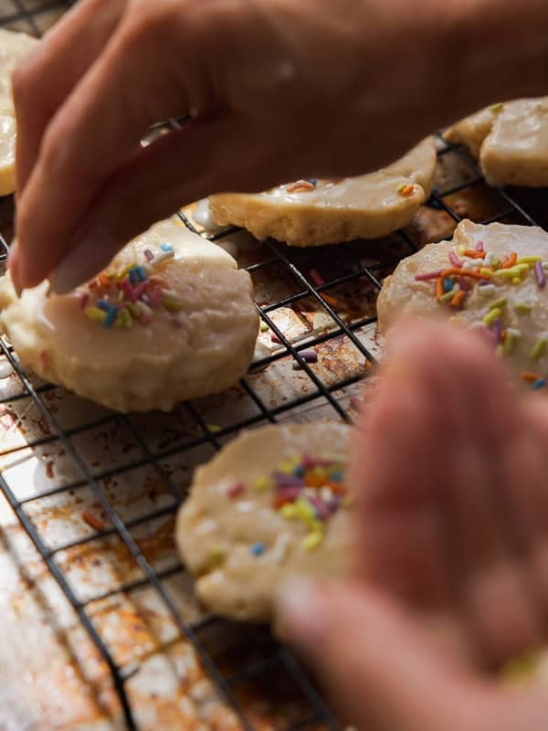Easy Vegan Lemon Cookies Recipe (Soft and Lemony!) 30 A close-up of hands decorating round lemon cookies with colorful sprinkles on a wire cooling rack. The cookies are covered in icing, and the background shows a baking tray underneath the rack.