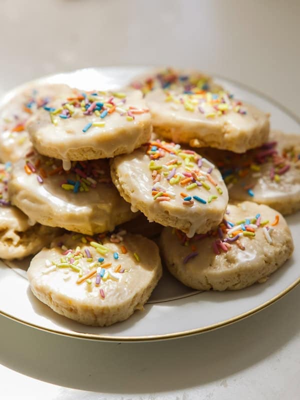 Easy Vegan Lemon Cookies Recipe (Soft and Lemony!) 31 A plate of round, glazed lemon cookies topped with colorful sprinkles, stacked in a pile on a white surface with sunlight shining on them.