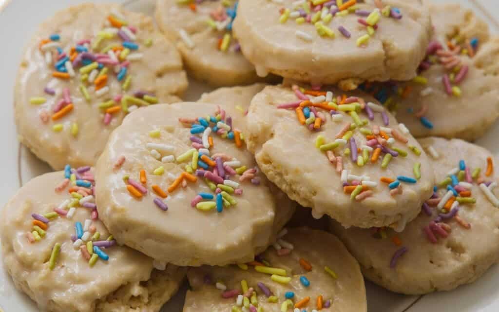 Easy Vegan Lemon Cookies Recipe (Soft and Lemony!) 1 A plate of frosted lemon cookies topped with colorful rainbow sprinkles, arranged in a pile. The cookies have a soft texture and a glossy layer of icing.