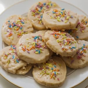 Easy Vegan Lemon Cookies Recipe (Soft and Lemony!) 33 A plate of round lemon cookies topped with white icing and colorful rainbow sprinkles, arranged in a pile on a white and gold-rimmed plate.