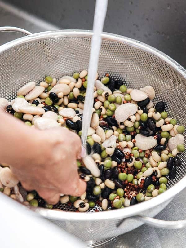 Gut Healthy Korean Multigrain Rice - Japgokbap (잡곡밥) 13 A person rinses a mix of dried beans and peas for Japgokbap, or Korean multigrain rice, in a metal colander under running water, preparing a gut healthy meal in the kitchen sink.
