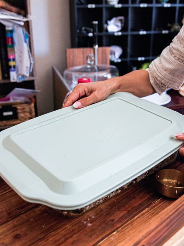 Gut Healthy Korean Multigrain Rice - Japgokbap (잡곡밥) 17 A person places a light green plastic lid on a rectangular baking dish of Korean Multigrain Rice (Japgokbap) on a wooden table, with kitchen items and shelves visible in the background for a gut healthy meal.