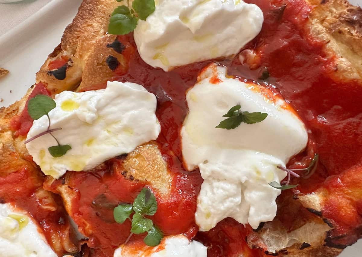 Close-up of a rustic pizza topped with creamy burrata cheese, vibrant Mutti tomato sauce, microgreens, and olive oil on a golden, slightly charred crust—reminiscent of flavors from Parma, Italy.