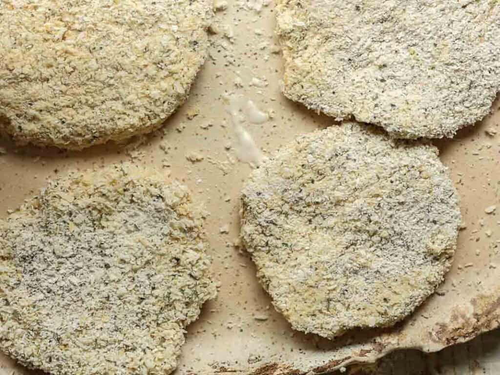 Easy & Delicious Hi-Protein Chickpea Cutlets 8 Four breaded, uncooked chickpea cutlets are arranged on a light-colored surface, with some crumbs scattered around them. The patties appear to be coated in seasoned breadcrumbs, ready for baking or frying.