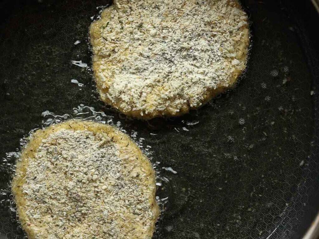 Easy & Delicious Hi-Protein Chickpea Cutlets 9 Two breaded chickpea cutlets are frying in oil in a dark nonstick pan, with visible bubbles forming around the edges of the patties.