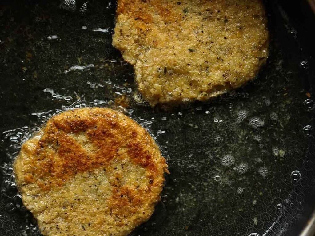 Easy & Delicious Hi-Protein Chickpea Cutlets 10 Two golden-brown chickpea cutlets are frying in oil in a dark pan, with visible bubbles around the edges, indicating they are being cooked.