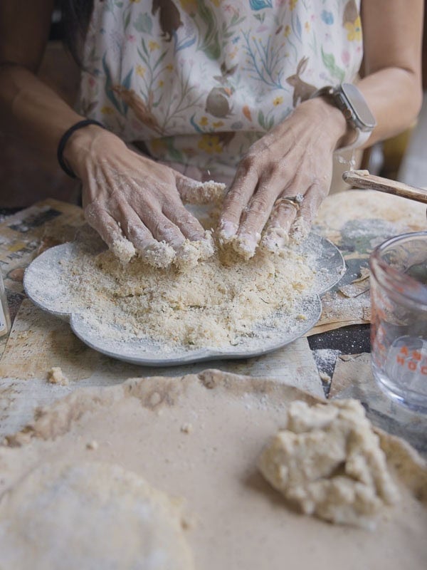 Easy & Delicious Hi-Protein Chickpea Cutlets 7 A person wearing a patterned apron mixes flour and dough for chickpea cutlets with their hands on a plate, surrounded by baking tools and ingredients on a kitchen counter.