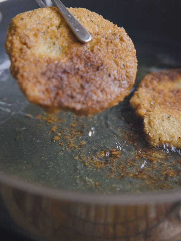 Easy & Delicious Hi-Protein Chickpea Cutlets 11 A chickpea cutlet is being lifted from a frying pan with oil using a metal utensil, while another cutlet remains in the pan. The chickpea cutlets are golden brown and crispy.