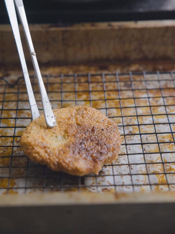 Easy & Delicious Hi-Protein Chickpea Cutlets 12 A pair of metal tongs holds a golden-brown, breaded chickpea cutlet on a wire rack set over a baking tray, likely cooling after frying.