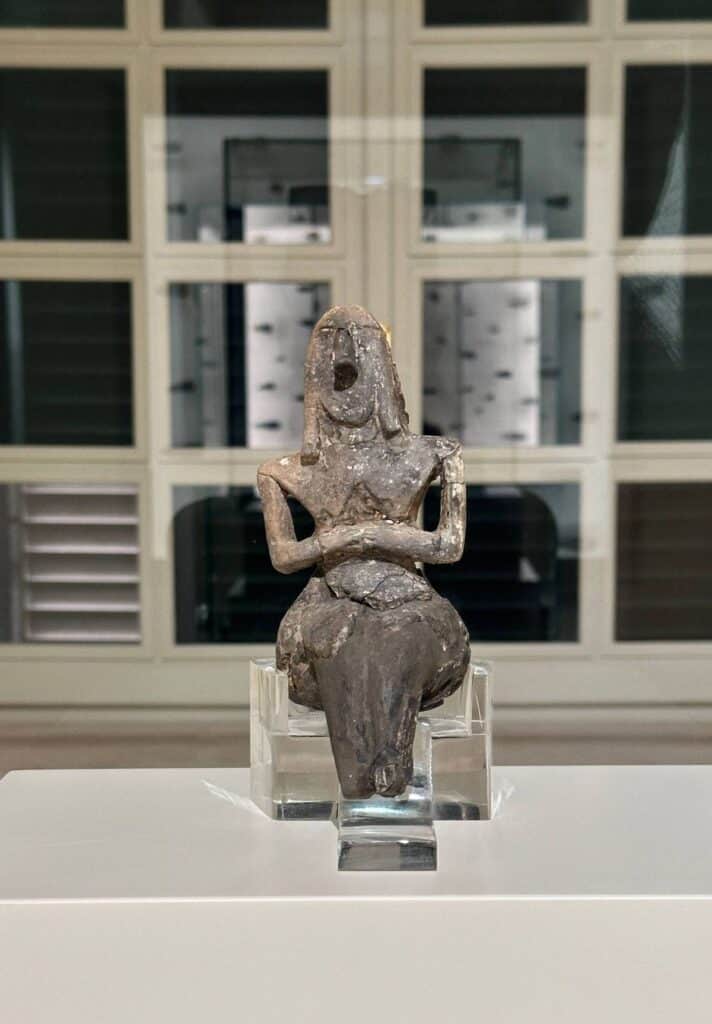 A stone figurine of a seated woman with pronounced hips and crossed arms is displayed on a clear stand in a museum setting.