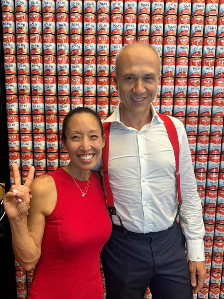 Two people smiling—one woman in a red sleeveless top making a peace sign, and one man in a white shirt with red suspenders—stand before a wall stacked with Mutti tomato cans, celebrating the taste of Parma’s finest.