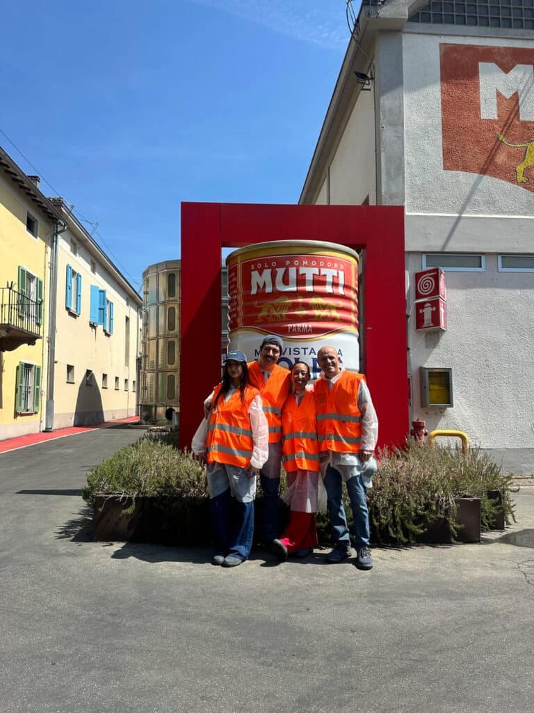 Da Vinci & Mutti: A Visit to Parma, Italy. 15 Three people in orange safety vests and protective gear stand smiling in front of a giant Mutti tomato can display outside a building in Parma, Italy, on a sunny day.