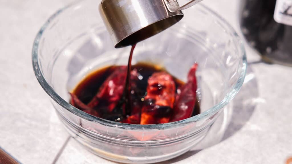 BEST Baby Bok Choy Kimchi Recipe - Easy & Delicious 22 A metal measuring cup pours dark liquid over dried red chilies in a clear glass bowl, sitting on a light-colored surface—an essential step when preparing flavorful bok choy kimchi.