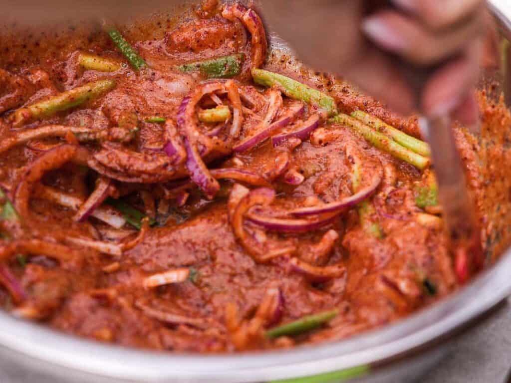 BEST Baby Bok Choy Kimchi Recipe - Easy & Delicious 41 A close-up of a bowl containing sliced red onions and green vegetables, including bok choy kimchi, mixed with a red, spicy marinade, being stirred by a person’s hand.