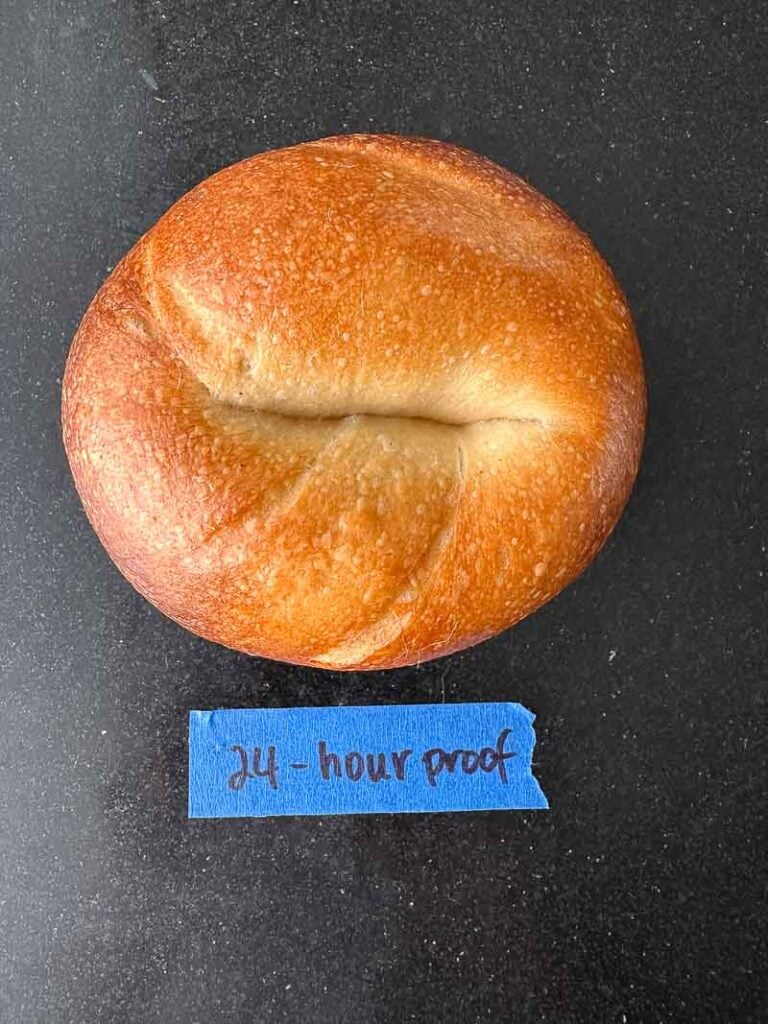 How to Make Delicious NY Style Bagels - Step by Step Guide. 80 A round, golden-brown bread roll, reminiscent of classic NY style bagels, sits on a dark surface next to a piece of blue tape labeled 24-hour KC handwritten in blue ink.