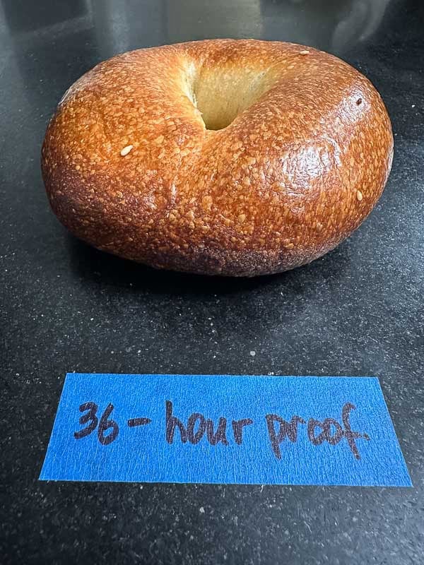 How to Make Delicious NY Style Bagels - Step by Step Guide. 83 A single NY style bagel with a dark, crisp crust sits on a black surface above a piece of blue tape labeled 36-hour proof handwritten in black marker.