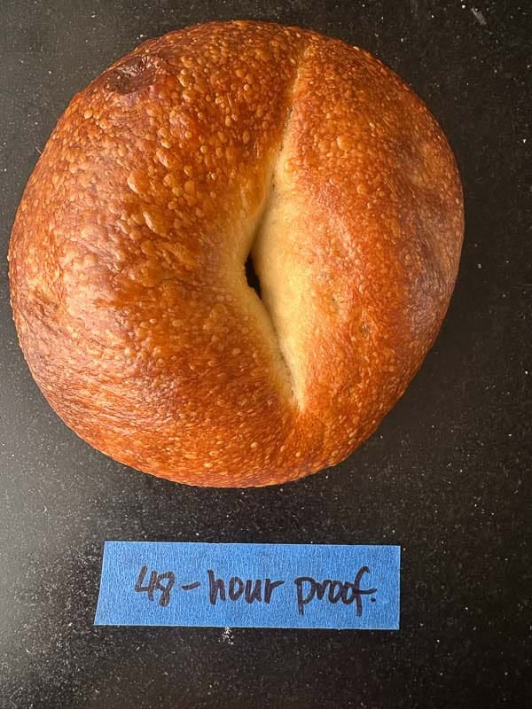 How to Make Delicious NY Style Bagels - Step by Step Guide. 84 A golden brown, NY style bagel sits on a dark surface above a blue label that reads 48-hour proof in black marker.