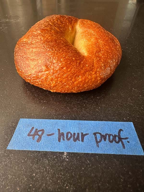 How to Make Delicious NY Style Bagels - Step by Step Guide. 85 A single, golden-brown NY style bagel sits on a dark surface behind a blue label that reads 48-hour proof.