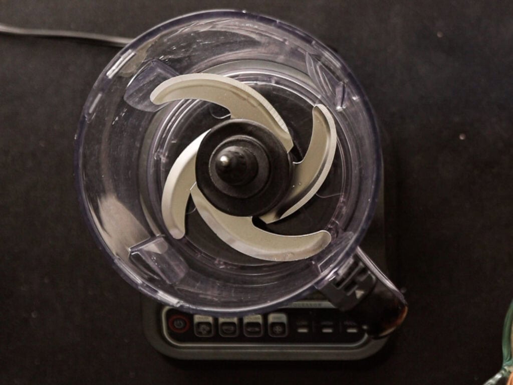 How to Make Delicious NY Style Bagels - Step by Step Guide. 9 Top-down view of an empty food processor with sharp metal blades inside, placed on a dark surface with some buttons visible on its base—perfect for preparing dough to make authentic NY style bagels at home.