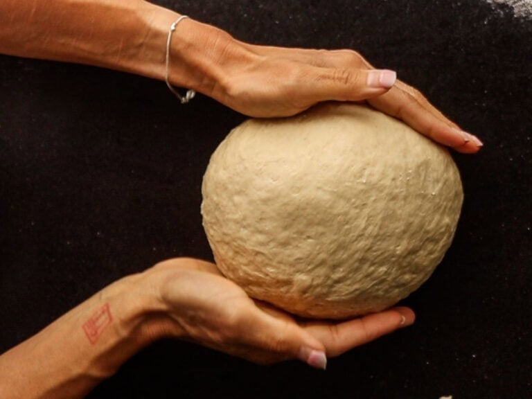 Two hands cradle a smooth ball of dough against a dark surface, with a silver bracelet on one wrist and a faint red stamp visible on the arm—perfectly capturing the artisanal process behind making authentic NY style bagels.