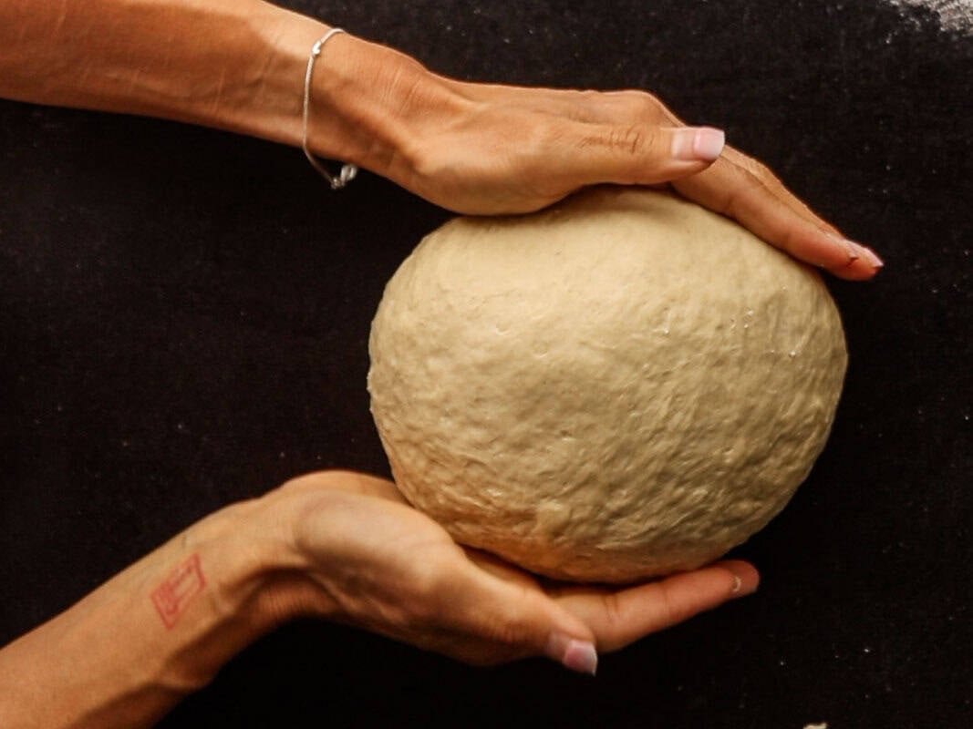 How My Bagels Triggered The Sexist In Me. 1 Two hands cradle a smooth ball of dough against a dark surface, with a silver bracelet on one wrist and a faint red stamp visible on the arm—perfectly capturing the artisanal process behind making authentic NY style bagels.
