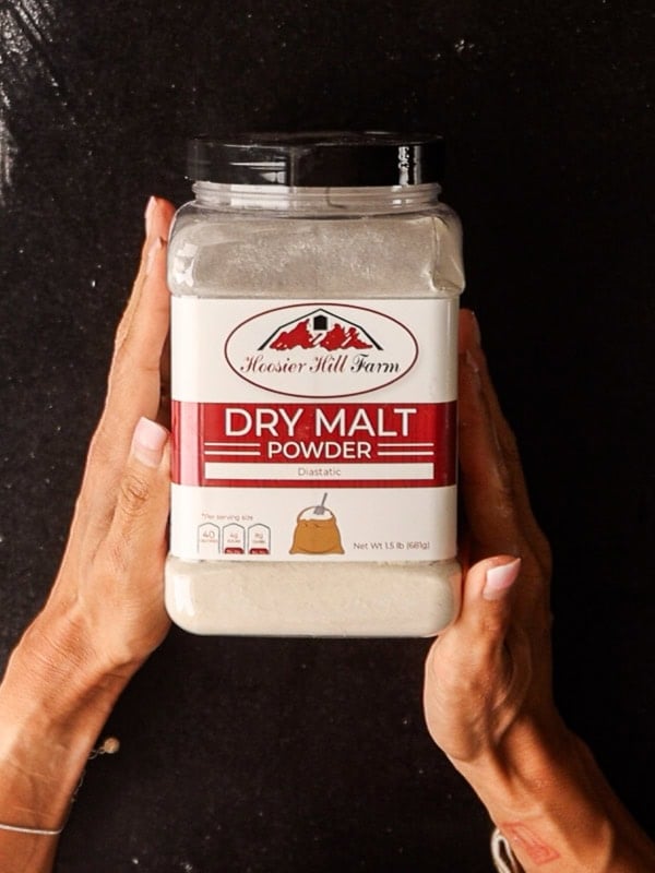 How to Make Delicious NY Style Bagels - Step by Step Guide. 14 Two hands hold a container of Hoosier Hill Farm Dry Malt Powder with a red and white label against a black background—perfect for baking authentic NY style bagels at home.