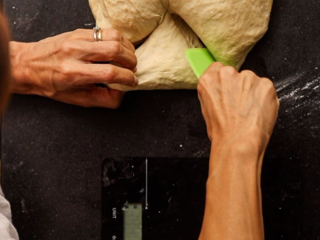 How to Make Delicious NY Style Bagels - Step by Step Guide. 24 Hands work with dough on a dark surface, using a green dough scraper to divide it, preparing classic NY style bagels. A digital kitchen scale is partially visible underneath, and some flour is scattered nearby.