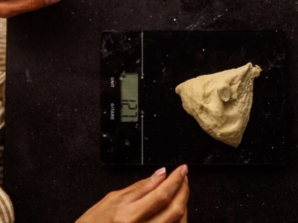 How to Make Delicious NY Style Bagels - Step by Step Guide. 25 A piece of dough weighing 127 grams sits on a digital kitchen scale, ready to be shaped into classic NY style bagels, with a person’s hands visible on a dark countertop.