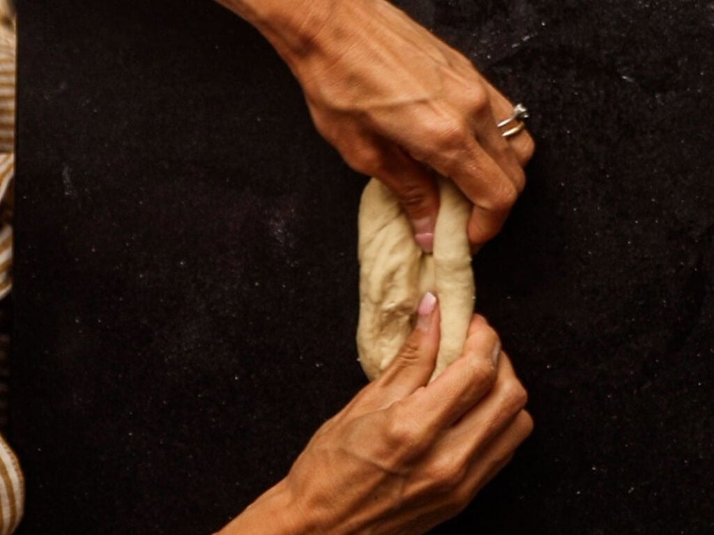 How to Make Delicious NY Style Bagels - Step by Step Guide. 26 Two hands are folding a piece of dough on a dark surface, preparing it for baking authentic NY style bagels. One hand has a ring.